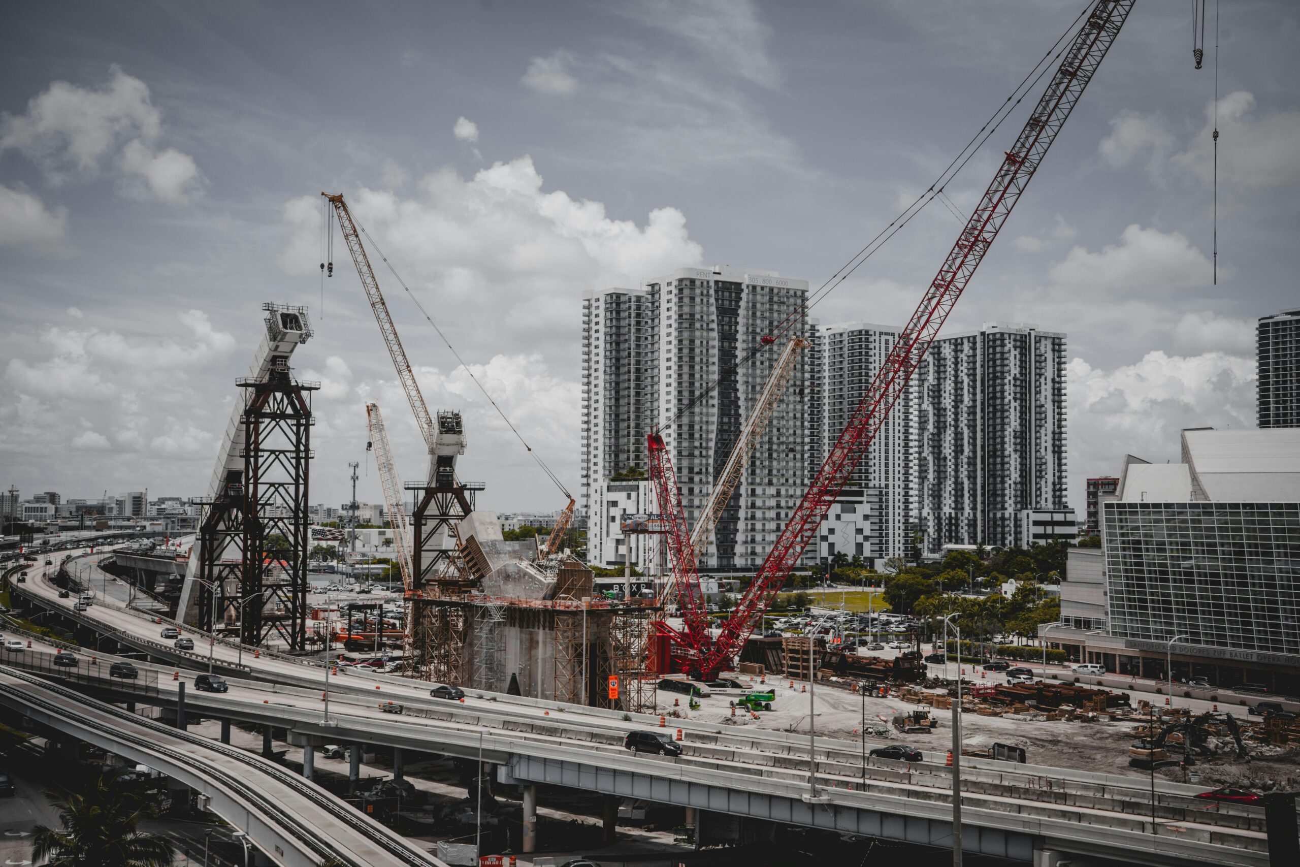 A cityscape featuring high-rise buildings and a construction site with cranes under a cloudy sky.